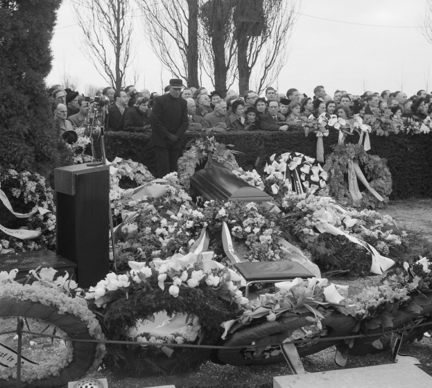 Schwarzer und weißer Trauerzug mit einem Mann in einer Mütze, der auf einem Podium spricht und von Blumen, Stühlen und einer Menge umgeben ist, vor Bäumen und einem klaren Himmel.