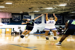 Eine Gruppe von Menschen, die Basketball in einer Turnhalle spielt, mit Zuschauern im Hintergrund, Bannern und einer Flagge an der Wand und einer Decke mit Stangen und Lampen oben.