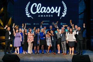 Eine Gruppe von Menschen auf einer Bühne, die strahlen und Auszeichnungen halten, mit Lautsprechern unten und einem Bildschirm im Hintergrund, der "Classy Awards 2019" anzeigt.