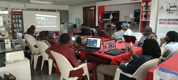 Gruppe von Menschen, die um einen Tisch mit Laptops, Flaschen und anderen Gegenständen sitzen, während eines digitalen Marketing-Workshops, mit einem Banner, einem Bildschirm, einer Tür und Deckenleuchten im Hintergrund.