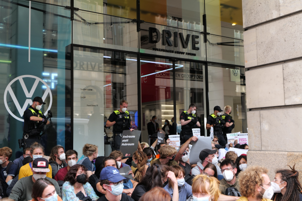 Eine große Gruppe von Menschen mit Masken und Schildern vor einem Glasgebäude mit Text, einige tragen Mützen und halten Kameras, was auf eine Demonstration in Melbourne, Australien, hindeutet.