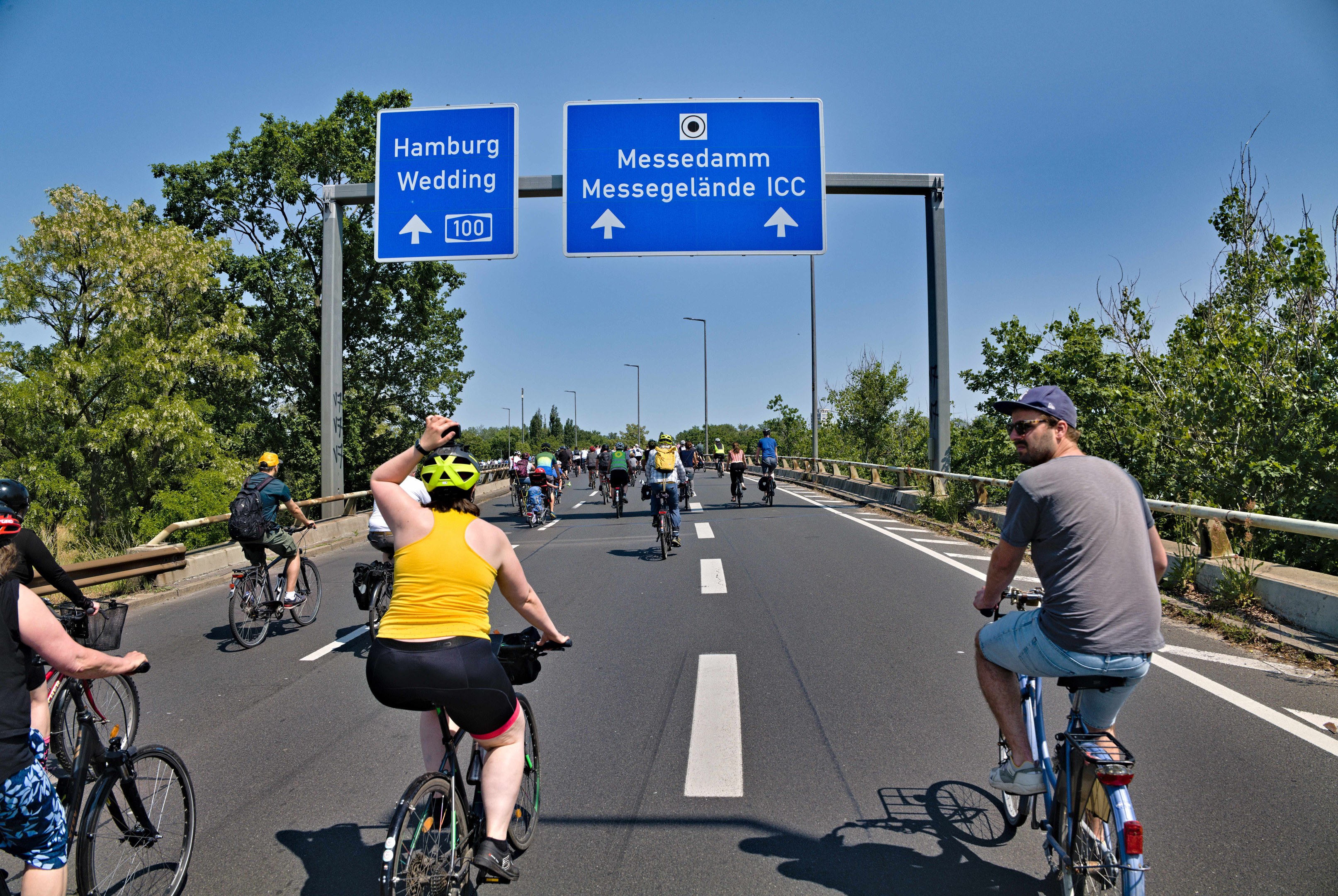 Gruppe von Radfahrern mit Helmen auf einer Straße mit Geländer und Bäumen auf beiden Seiten, Laternen im Hintergrund und einem klaren blauen Himmel, mit einer Tafel, die eine Radtour in Hamburg anzeigt.
