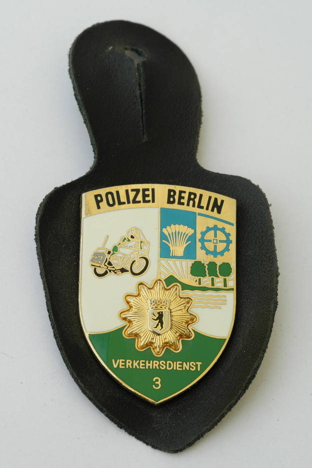 Ein schildförmiges Polizeibadged mit "Polizei Berlin" in fetter schwarzer Schrift, das ein weißes Stern auf blauem Hintergrund und einen weißen Rand hat und auf einer Oberfläche liegt.