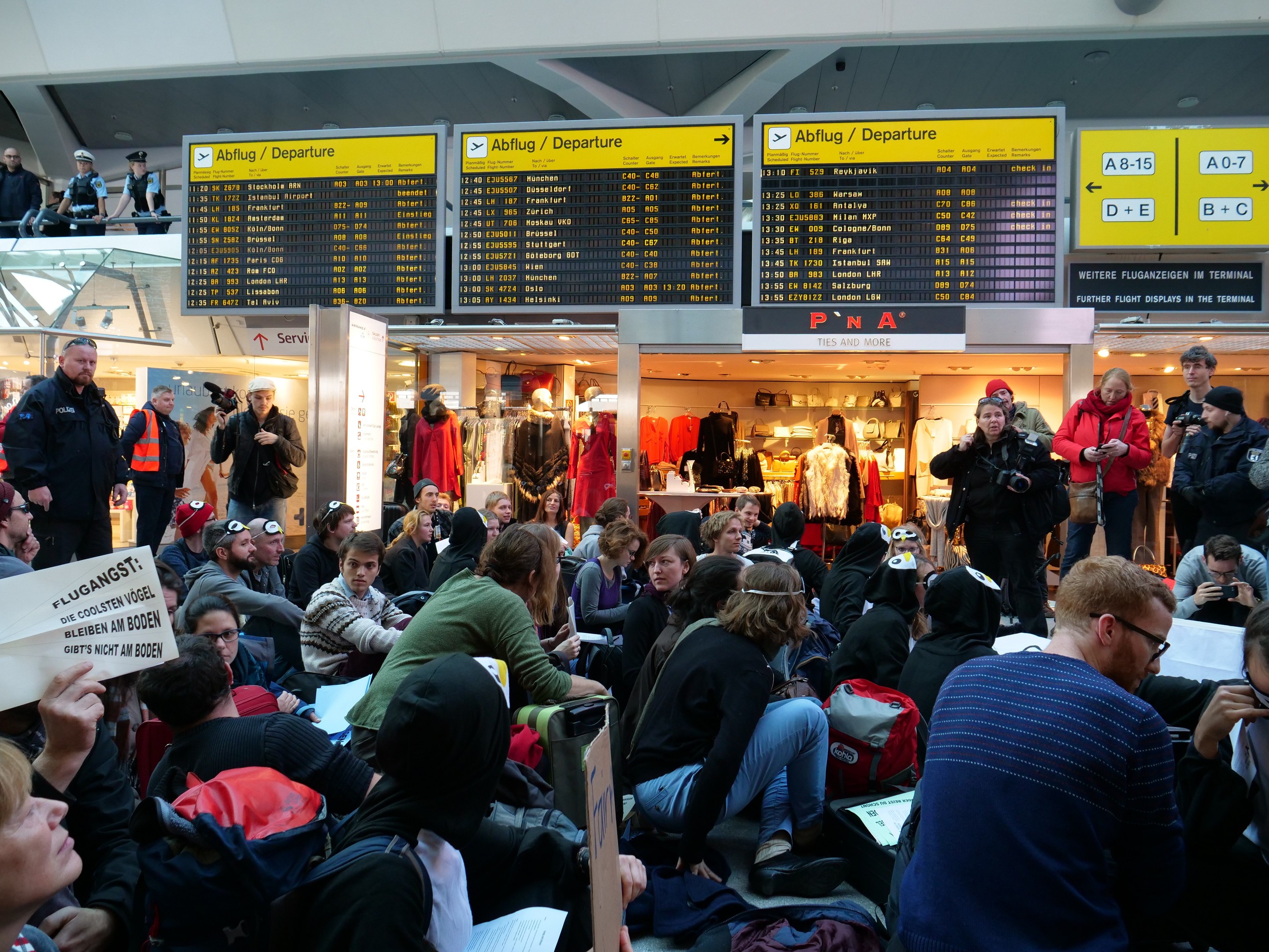Eine große Gruppe von Menschen an einem Flughafen, einige sitzen mit Taschen und Papieren, andere stehen, mit Texttafeln, Schaufensterpuppen in Kleidern und Deckenbeleuchtung im Hintergrund, was auf eine Protestveranstaltung hindeutet.