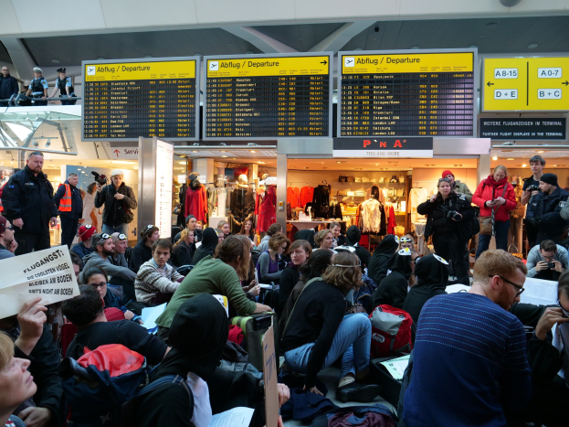 Eine große Gruppe von Menschen an einem Flughafen, einige sitzen mit Taschen und Papieren, andere stehen, mit Texttafeln, Schaufensterpuppen in Kleidern und Deckenbeleuchtung im Hintergrund, was auf eine Protestveranstaltung hindeutet.