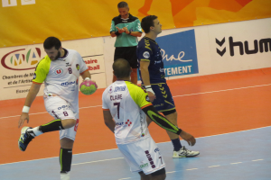 Eine Gruppe von Männern spielt Handball auf einem Platz, wobei einer einen Ball hält und im Hintergrund Schilder mit Text zu sehen sind.