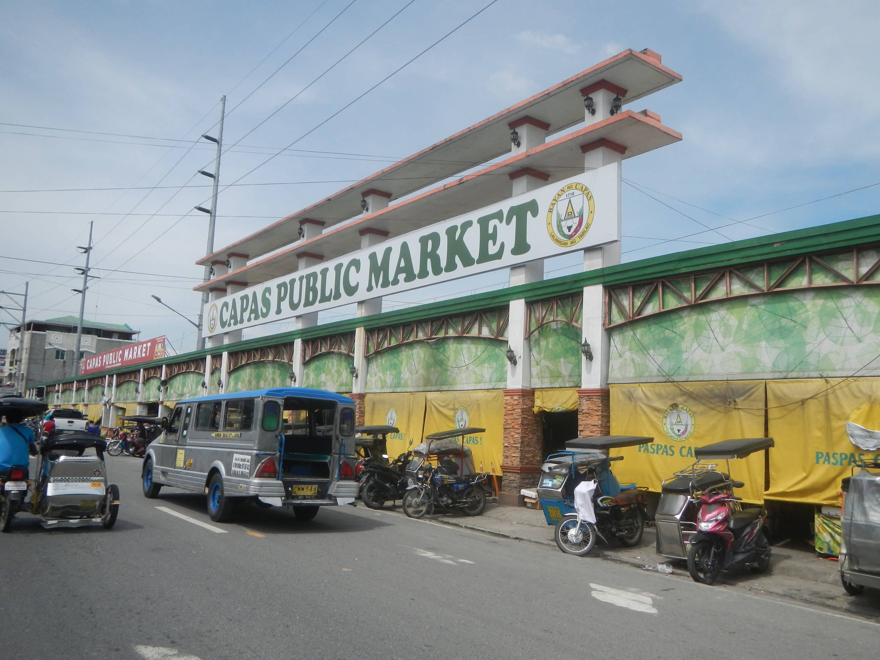 Belebte Stadtstraße mit Autos, Motorrädern und Rikschas vor einem Gebäude mit der Aufschrift "Capas Public Market", Strommasten, Laternenmasten und Gebäude im Hintergrund bei bewölktem Himmel.