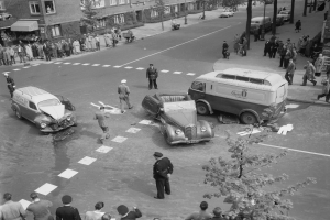 Schwarz-weißes Bild einer Autounfall-Szene an einer Straßenkreuzung mit mehreren Fahrzeugen, darunter ein Lieferwagen, und einer Gruppe von Menschen in der Nähe, mit Gebäuden, Bäumen, Pfählen und Texttafeln im Hintergrund.