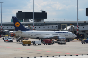 Ein Lufthansa Airbus A320-200-Flugzeug steht auf dem Frankfurter Flughafen, mit Fahrzeugen und Wagen auf der Straße davor, einem Gebäude, Laternenpfählen und einem klaren blauen Himmel im Hintergrund.