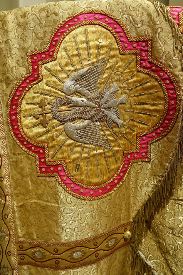 Nahaufnahme eines Priestergewandes mit aufwendiger goldener und roter Stickerei vor einem einfarbigen Wandhintergrund.