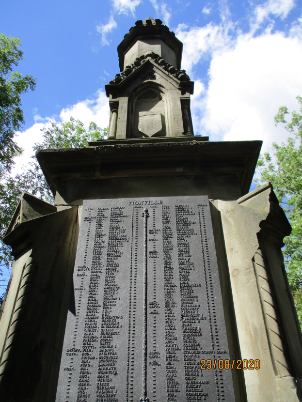 Ein steinernes Denkmal mit einem zentralen Uhrenturm, umgeben von Bäumen, mit eingravierten Texten, die wahrscheinlich Kriegsopfer gedenken, unter einem bewölkten Himmel.