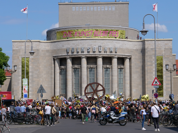 Eine große Gruppe von Menschen versammelt sich vor einem Gebäude mit Säulen und Text, umgeben von Straßenlaternen, Fahrzeugen und Bäumen, während einer Demonstration in Berlin, Deutschland, unter einem bewölkten Himmel.
