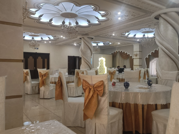 Großer Raum mit Tischen und Stühlen, die für eine Hochzeitsfeier arrangiert sind, Stühle mit Tüchern drapiert und mit dekorativen Accessoires versehen, Wände mit Türen und Vorhängen verziert, Decke mit Lichtern und Kronleuchtern geschmückt.