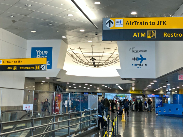 Eine Gruppe von Menschen geht durch eine Flughafenhalle mit einer Rolltreppe auf der linken Seite, Schilder an den Wänden mit Text, Lampen an der Decke und ein paar Menschen, die auf dem Boden stehen; ein Schild trägt den Text "Airtrain nach JFK".
