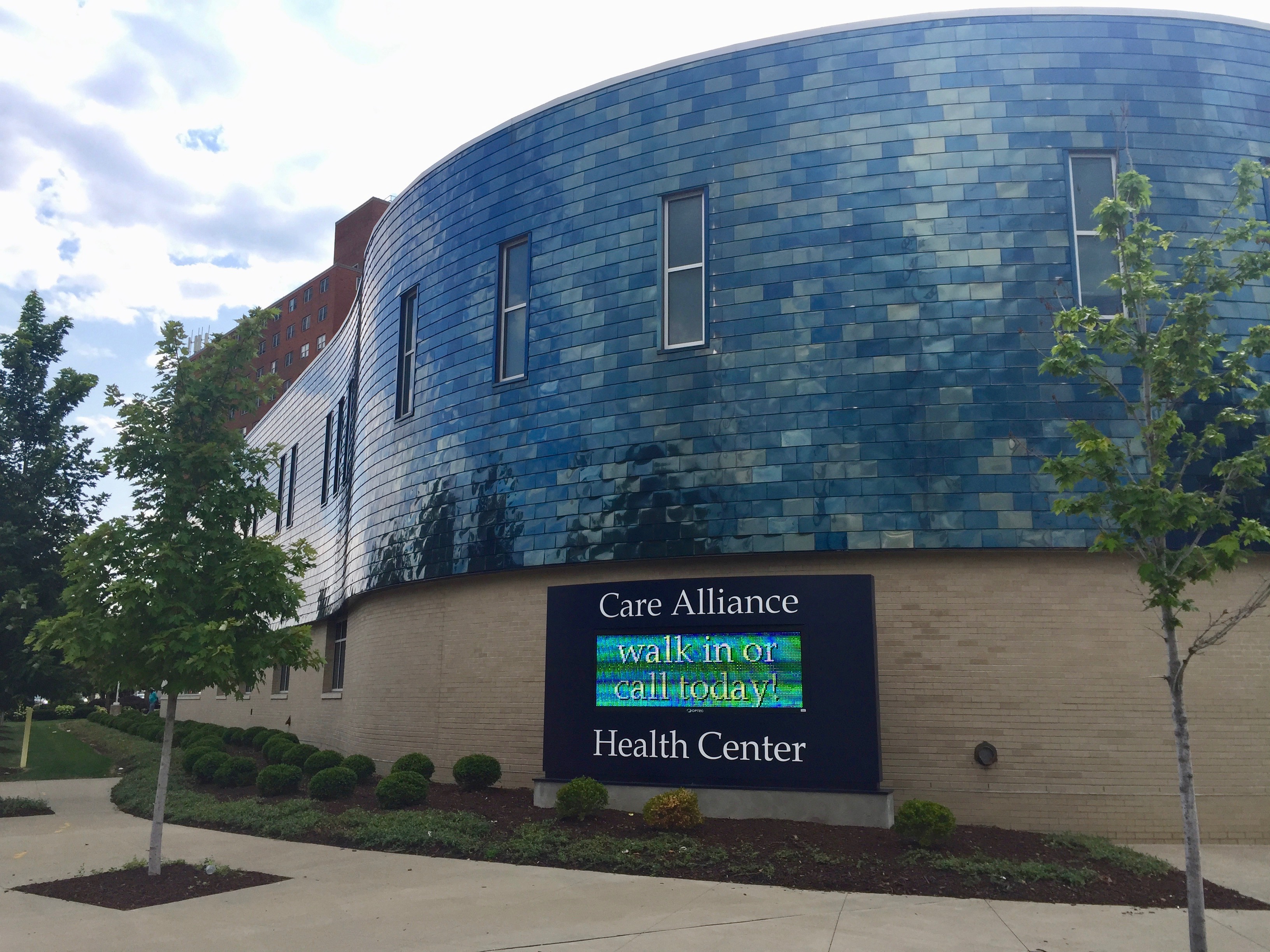 Ein Gebäude mit Fenstern und einem Schild, umgeben von Bäumen, Pflanzen und Gras, mit einem Weg, der darauf führt, unter einem bewölkten Himmel.