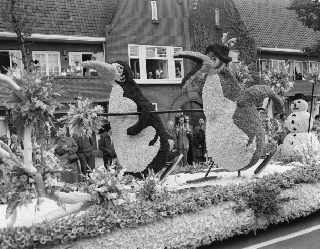 Schwarzes und weißes Foto eines Paradewagens, der mit Blumen und Pinguinen geschmückt ist, mit Menschen, Bäumen und Gebäuden mit Fenstern im Hintergrund.
