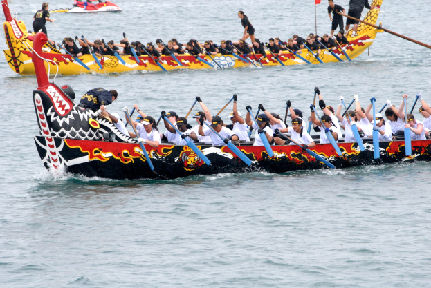 Eine Gruppe von Menschen in einem Drachenboot, das auf dem Wasser fährt und Paddel hält, mit einem anderen Boot und einer Fahne im Hintergrund.
