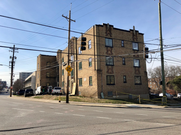 Großes Ziegelgebäude an einer Straßenecke mit Strommasten, Ampeln, Fahrzeugen, Bäumen und Wolken im Hintergrund, das in Wohnungen umgebaut wurde.