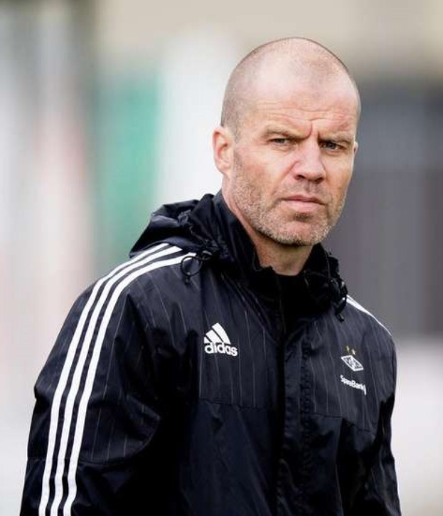 Kahlkopfiger Trainer in marineblauem Jackett mit weißen Streifen und weißem Kragen auf einem Fuballfeld stehend, Hände verschränkt, mit ernster Miene und unscharfem Hintergrund.