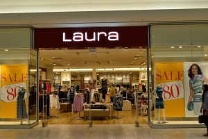 Innenansicht eines Laura Bekleidungsgeschäfts in einem Einkaufszentrum mit Schaufensterpuppen in Kleidern, hängenden Kleidungsstücken, Informationsschildern, Deckenleuchten und einer Person mit einer Tasche.