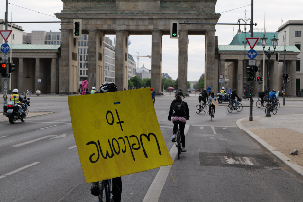 Eine Gruppe von Radfahrern fährt am Brandenburger Tor in Berlin vorbei, einer hält ein gelbes Schild, mit Laternenmasten, Verkehrsampeln, Gebäuden und Bäumen im Hintergrund und einem klaren Himmel.