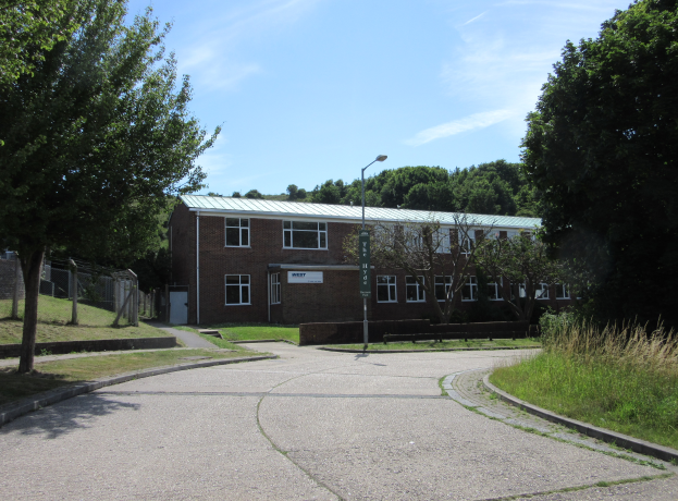 Ein Schulgebäude mit Fenstern befindet sich inmitten eines Parks, umgeben von saftigem Grün, mit einem Straßenschild, einem Schild, einer Baumgruppe, einem Zaun und einem bewölkten Himmel.