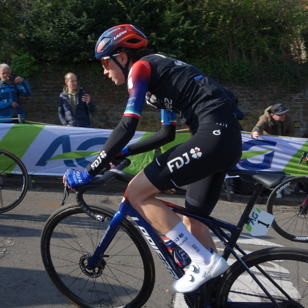 Eine professionelle Radrennfahrerin, Katie Archibald, fährt in Helm, Brille, Handschuhen und Schuhen ein Fahrrad eine Straße entlang neben einer Menge, mit einem Banner, einer Wand, Bäumen und einem Gebäude im Hintergrund.