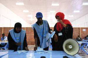 Eine Gruppe von Menschen steht um einen Tisch mit einem Megafon, Papieren und anderen Gegenständen herum, in einem Raum, der wie eine Wahllokal aussieht, mit mehr Menschen im Hintergrund unter einer Decke mit Lampen.