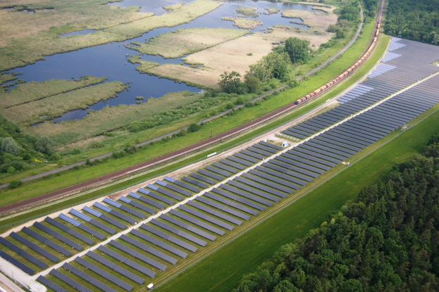 Luftaufnahme einer Solarfarms mit Solarpanelen in einem Feld, umgeben von Bäumen, Gras, Pflanzen und Wasser, mit einem Zug auf einer nähergelegenen Eisenbahnschiene.