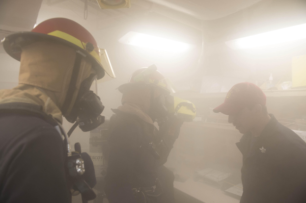 Feuerwehrleute in einem rauchgefüllten Raum während einer Übung, einer hält eine Kamera, mit Hintergrundlichtern und Equipment im Hintergrund.