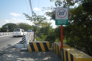 Ein Auto fährt auf einer Straße mit einem Tempolimit-Schild, Geländern und Bäumen auf beiden Seiten, unter einem bewölkten Himmel mit einem Wasserzeichen.