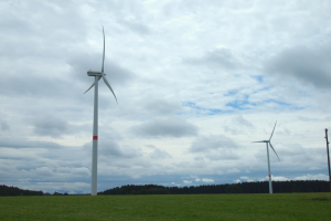Drei hohe, weiße Windräder auf einer grünen Wiese mit Bäumen und Wolken im Hintergrund.