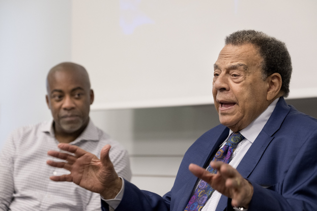 Zwei Männer sitzen nebeneinander vor einem Projektionsschirm, einer im Anzug und Krawatte, der andere in legerer Kleidung, beide mit ernsten Gesichtern.