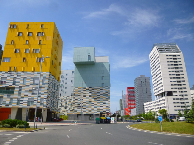 Eine Stadtstraße mit hohen Gebäuden, die als Teil der Universität Rotterdam gekennzeichnet sind, Straßenmasten, Straßenlaternen, Kraftfahrzeuge, Informationsschilder, Sträucher, Bäume und ein bewölkter Himmel.