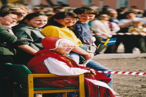 Alte Frau in einem Stuhl sitzend, die eine Menge ansieht, wobei einige Personen Brillen tragen und eine eine Flagge hölt; ein Band ist im Vordergrund und ein leicht unscharfer Gebäudehintergrund mit Fenstern.