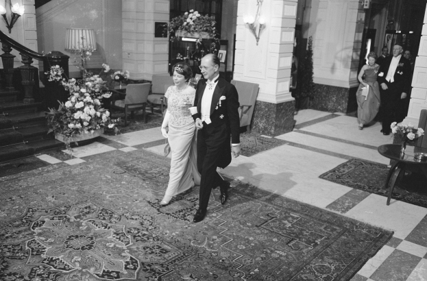 Schwarz-weiß-Foto einer Braut und eines Bräutigams, die durch die Hotellobby gehen und von Gästen umgeben sind, mit Blumengestecken, Möbeln und architektonischen Details.