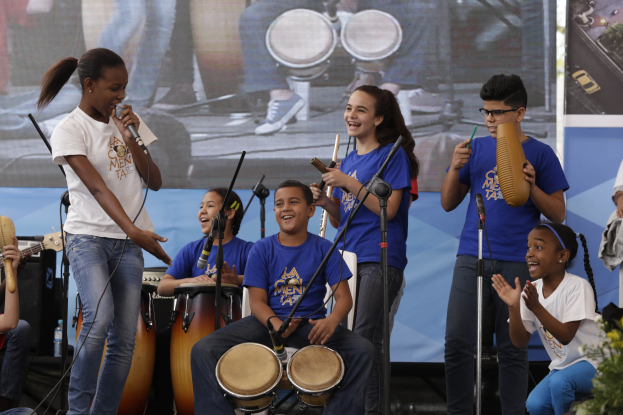 Kinder mit Musikinstrumenten auf einer Bühne mit einer Frau mit Mikrofon in der Mitte, umgeben von Mikrofonstativen, einem Banner im Hintergrund und Pflanzen auf der rechten Seite.
