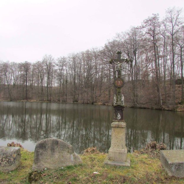 Ein Friedhof mit einem Kreuz in der Mitte eines Sees, umgeben von Bäumen, Grabsteinen und saftigem Grün, unter einem sichtbaren Himmel.