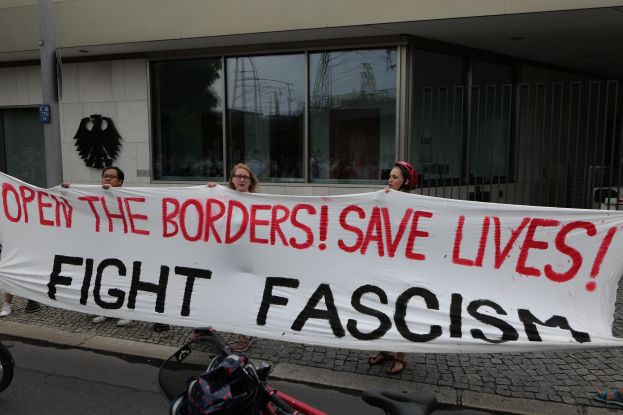 Eine Gruppe von Menschen hält ein Transparent mit der Aufschrift "Öffnet die Grenzen, rettet Leben, kämpft gegen Faschismus" vor einem Gebäude mit Glasfenstern und einem Pfahl, mit einem Fahrrad und einer Tasche auf der Straße.