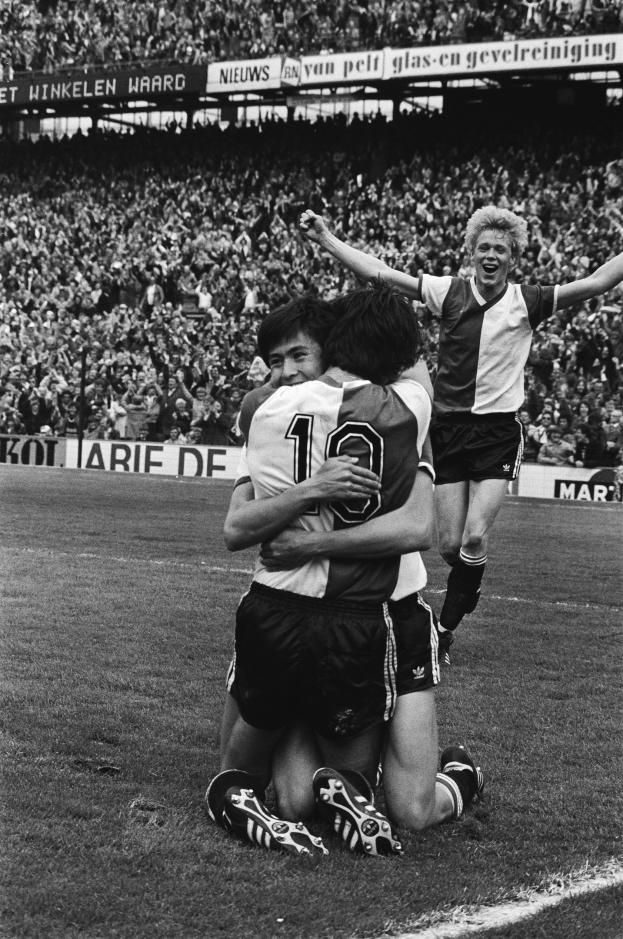 Drei Fußballer umarmen sich auf dem Boden nach einem Tor, mit jubelnder Menge und schwenkenden Fahnen in einem schwarz-weißen Stadion-Setting.