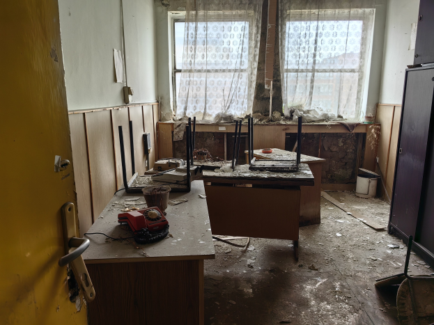 Leerer Raum mit einer Tür auf der linken Seite, einem Schrank auf der rechten Seite und einem Fenster mit Vorhängen im Hintergrund; ein Tisch in der Mitte hält verstreute Gegenstände und vermittelt den Eindruck eines verlassenen Büros.