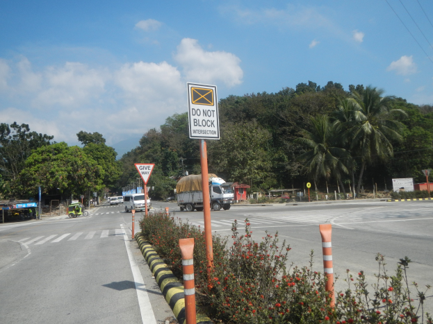Eine Straßensperre mit durchfahrenden Fahrzeugen, umgeben von Vegetation und einem "Do Not Block"-Schild unter einem bewölkten Himmel.