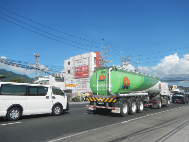 Ein gro├če Tanklastwagen fahrt auf einer Stra├če neben einem wei├če Van, mit elektrischen Masten, Geb├Ąuden, Plakatw├Ąnden, B├Ąumen, H├Ąngen und einem bew├Âlktem Himmel im Hintergrund.