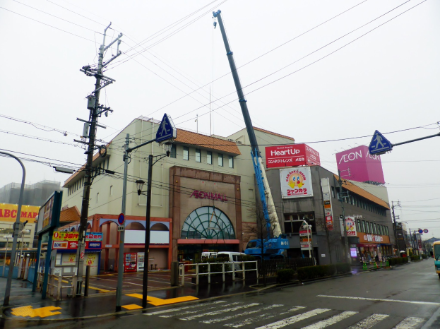 Eine Stadtstraße mit Fahrzeugen, Versorgungsmasten, Schildern, einem Kran, Gebäuden und einer großen Baustelle in der Mitte.