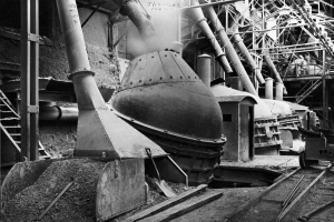 Ein Schwarz-Weiß-Foto einer großen Maschine in einer Fabrik, umgeben von verschiedenen Maschinen, Pfählen, Treppen, Geländern und anderen Gegenständen, mit Eisenbahnschienen unten.