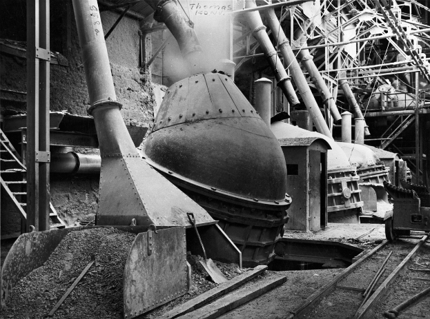 Ein Schwarz-Weiß-Foto einer großen Maschine in einer Fabrik, umgeben von verschiedenen Maschinen, Pfählen, Treppen, Geländern und anderen Gegenständen, mit Eisenbahnschienen unten.