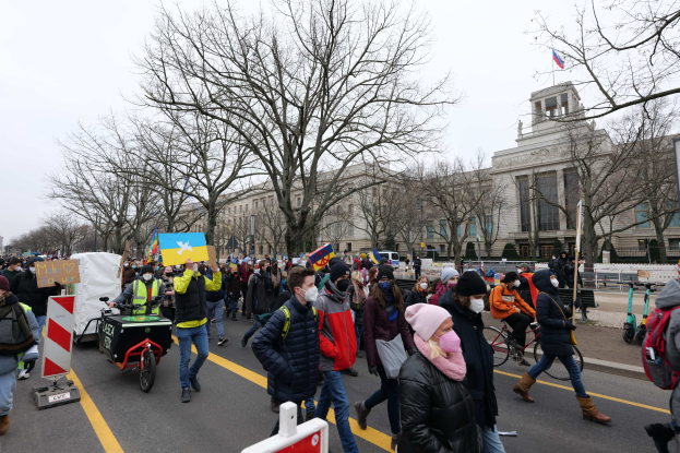 Eine große Gruppe von Menschen marschiert bei einer Demonstration auf einer Straße in Washington, D.C., mit Schildern und Fahrrädern, mit Bäumen und einem klaren blauen Himmel im Hintergrund.
