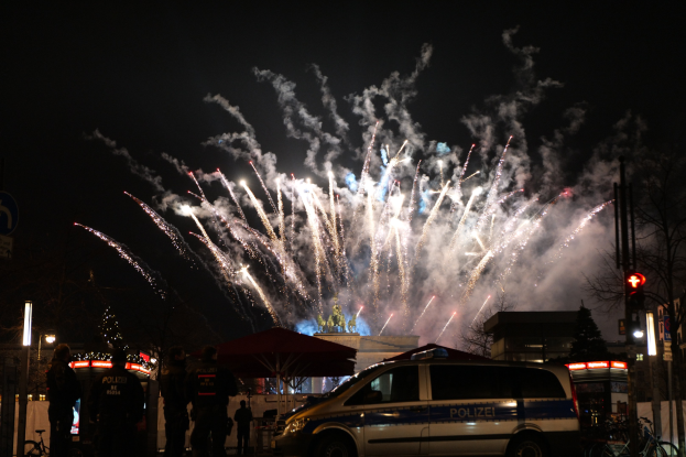 Ein Polizeiwagen vor einer Menschenmenge bei einer Silvesterfeier in Berlin geparkt, mit farbigen Feuerwerken, die den Himmel erhellen und die Stadtlichter