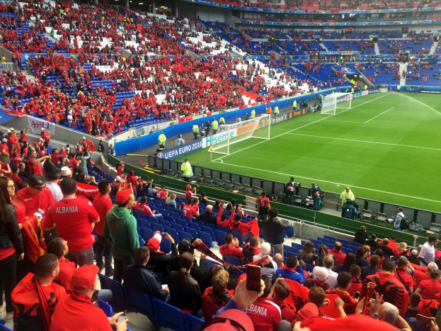Eine große Menschenmenge in einem Stadion schaut einem Fußballspiel zu, mit Menschen, die auf Stühlen sitzen und stehen, Fahnen, einem Torpfosten und Deckenleuchten.