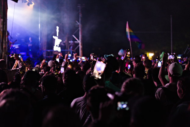 Eine Menge bei einem Konzert mit hochgehaltenen Handys, beleuchtet von Lichtern, umgeben von Pfählen und einer Fahne, während Musiker auf der Bühne spielen.